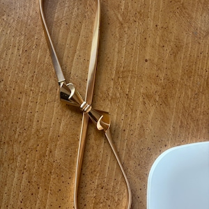 May include: A gold-toned metal belt featuring a flexible, coiled band and a central decorative bow-shaped clasp. The belt has a smooth, polished finish and a continuous, adjustable loop design that connects at the front with the bow element.