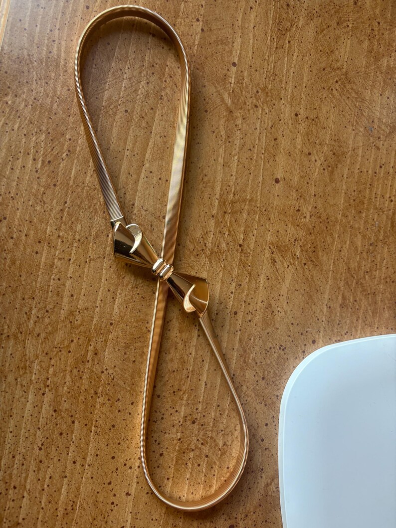 May include: A gold-toned metal belt featuring a flexible, coiled band and a central decorative bow-shaped clasp. The belt has a smooth, polished finish and a continuous, adjustable loop design that connects at the front with the bow element.