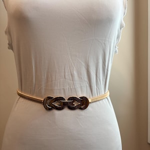 May include: A thin, gold-toned elastic belt featuring a central decorative buckle composed of two interlocking, polished metal loops with a textured, dark brown pattern. The belt is worn around the waist over a plain, sleeveless white top.