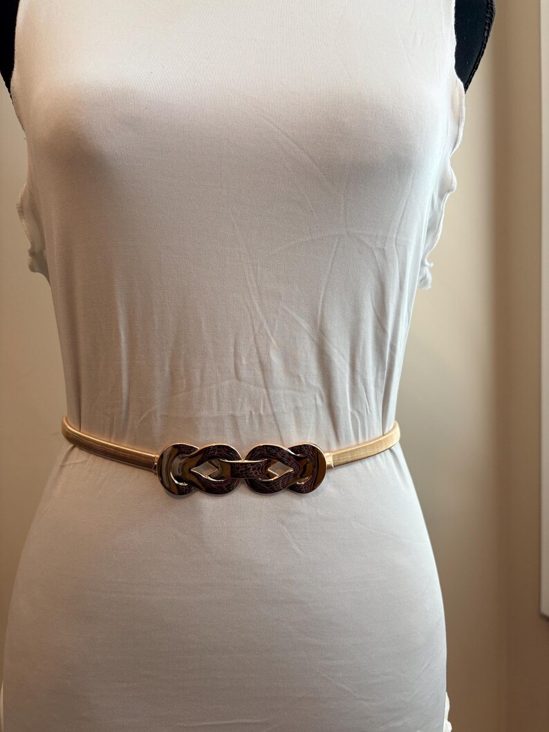 May include: A thin, gold-toned elastic belt featuring a central decorative buckle composed of two interlocking, polished metal loops with a textured, dark brown pattern. The belt is worn around the waist over a plain, sleeveless white top.