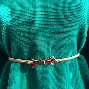 May include: A thin, metallic gold-toned belt featuring a central bow-shaped buckle. The belt is worn over a textured, vibrant green knit sweater, highlighting the contrast between the smooth, reflective metal finish and the fabric texture.