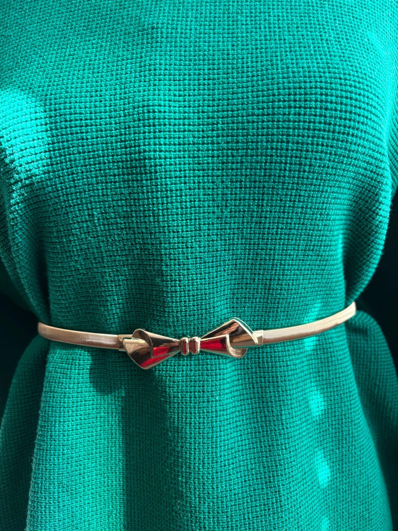 May include: A thin, metallic gold-toned belt featuring a central bow-shaped buckle. The belt is worn over a textured, vibrant green knit sweater, highlighting the contrast between the smooth, reflective metal finish and the fabric texture.