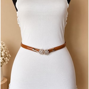 May include: A thin brown belt worn around a white sleeveless top. The belt features a metallic gold-tone clasp shaped like two adjacent flowers, each adorned with small clear crystals at the center and along the petals for a decorative finish.