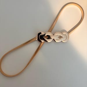 May include: A thin, gold-toned metallic belt featuring a flexible, coiled band and a central decorative buckle. The buckle consists of two interlocking, polished gold-colored loops that create a symmetrical, knot-like design in the center.