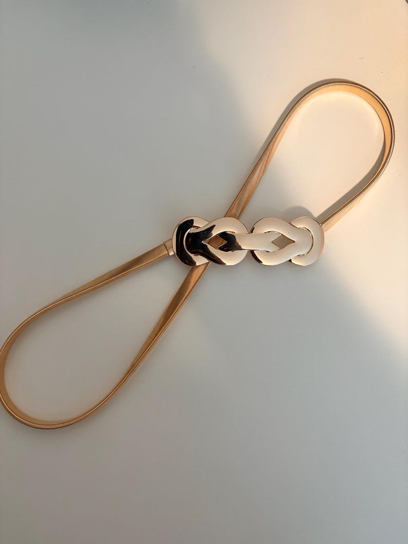 May include: A thin, gold-toned metallic belt featuring a flexible, coiled band and a central decorative buckle. The buckle consists of two interlocking, polished gold-colored loops that create a symmetrical, knot-like design in the center.