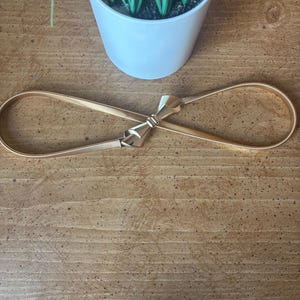 May include: A gold-toned metal belt featuring a flexible, thin band and a central bow-shaped clasp. The belt is designed with a smooth, reflective finish and a continuous loop structure that connects at the front with the decorative bow element.