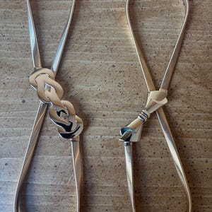 May include: Two gold-toned elastic belts with metallic finishes. The left belt features a decorative interlocking knot buckle, while the right belt displays a sleek, bow-shaped metal clasp. Both belts have thin, flexible bands with a fine ribbed texture.