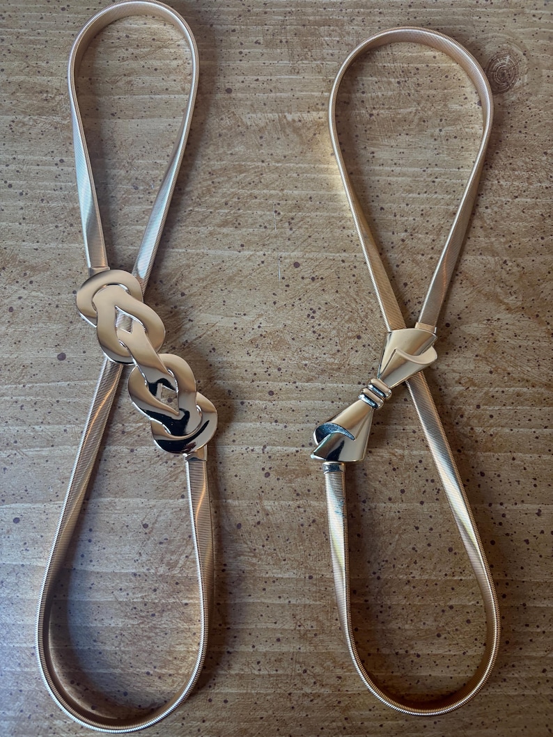 May include: Two gold-toned elastic belts with metallic finishes. The left belt features a decorative interlocking knot buckle, while the right belt displays a sleek, bow-shaped metal clasp. Both belts have thin, flexible bands with a fine ribbed texture.