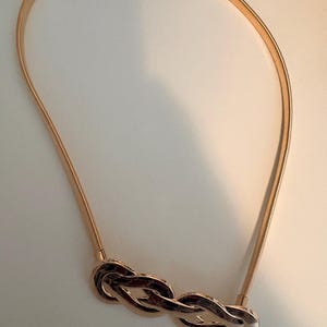 May include: A gold-toned metal belt featuring a flexible, thin, coiled band and a central decorative element composed of three interlocking, polished oval links that create a continuous, braided-style knot design in a shiny metallic finish.