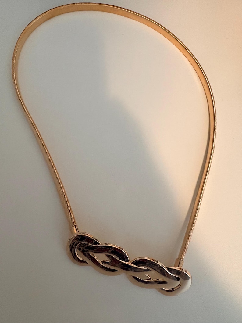 May include: A gold-toned metal belt featuring a flexible, thin, coiled band and a central decorative element composed of three interlocking, polished oval links that create a continuous, braided-style knot design in a shiny metallic finish.