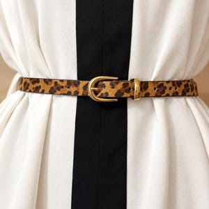 May include: A thin belt featuring a brown and black leopard print pattern. The belt is secured with a polished gold-tone metal buckle and a matching metal keeper loop, providing a functional closure for the adjustable strap design.