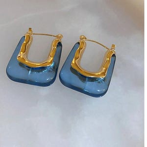 May include: A pair of blue and gold hoop earrings. The hoops are made of a clear blue material with a gold metal frame.