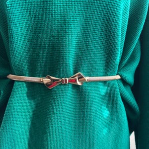 May include: A thin, metallic gold-toned belt featuring a central bow-shaped buckle, worn over a vibrant emerald green sweater with a textured, waffle-knit pattern. The belt is slender and polished, contrasting with the soft, knitted fabric.