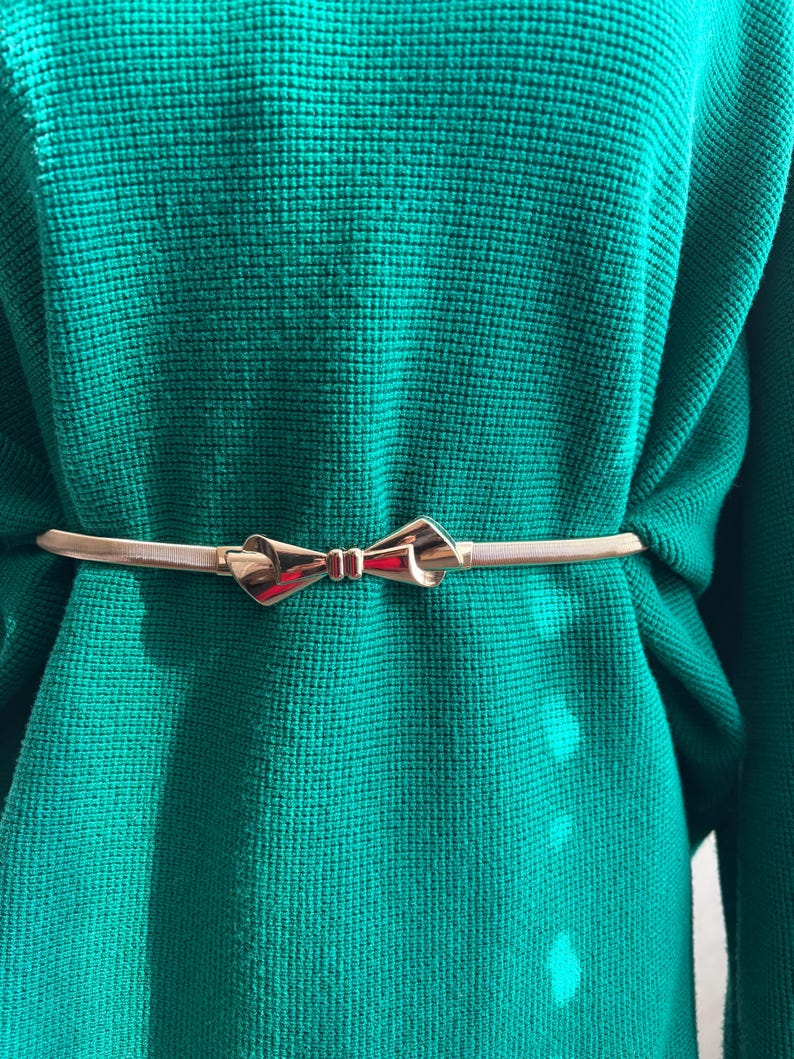 May include: A thin, metallic gold-toned belt featuring a central bow-shaped buckle, worn over a vibrant emerald green sweater with a textured, waffle-knit pattern. The belt is slender and polished, contrasting with the soft, knitted fabric.