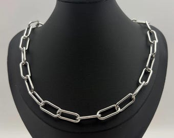May include: A silver-colored chain necklace with large, rectangular links. The necklace is displayed on a black mannequin bust. The links are evenly spaced and create a bold, modern look. The necklace is a statement piece.