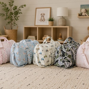 May include: Five cylindrical duffel bags with ruffled side panels and adjustable shoulder straps. The bags feature various patterns including pink floral, blue bird, cream tractor, green camouflage, and light pink line art designs.