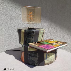 以下が含まれることがあります： 黒、白、オレンジの幾何学模様が施されたセラミック製のランプベース。ベースの上に、コイル状のフィラメント電球が付いた透明なガラス製のランプシェードが置かれている。カラフルな表紙の本がベースの上に置かれている。