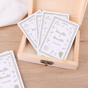 Peut inclure: Une boîte en bois clair avec un fermoir en métal contient plusieurs cartes blanches. Chaque carte présente des dessins au trait verts, les noms "Martha & Donald" et la date "12.22.2025". Les cartes ont une bordure décorative avec des illustrations sur le thème du mariage.