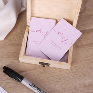May include: A wooden box with a hinged lid holds several light purple cards. Each card has the initials "m d" in a cursive font, along with the names "MARTHA & DONALD" and the date "Dec 22th 2025". A black Sharpie pen lies nearby.
