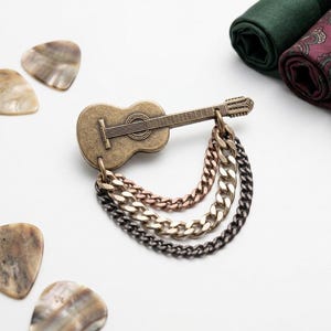 Könnte beinhalten: Eine bronzefarbene Gitarrenbrosche mit drei Ketten in Silber, Kupfer und Schwarz. Gitarrenplektren und gerollter Stoff in Grün, Burgunderrot und Braun mit Streifenmuster sind ebenfalls zu sehen.