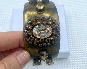 May include: A vintage-style brass cuff bracelet with an oval, clear crystal center stone surrounded by smaller brown and gold crystals. The bracelet has additional crystal accents and a tarnished finish.