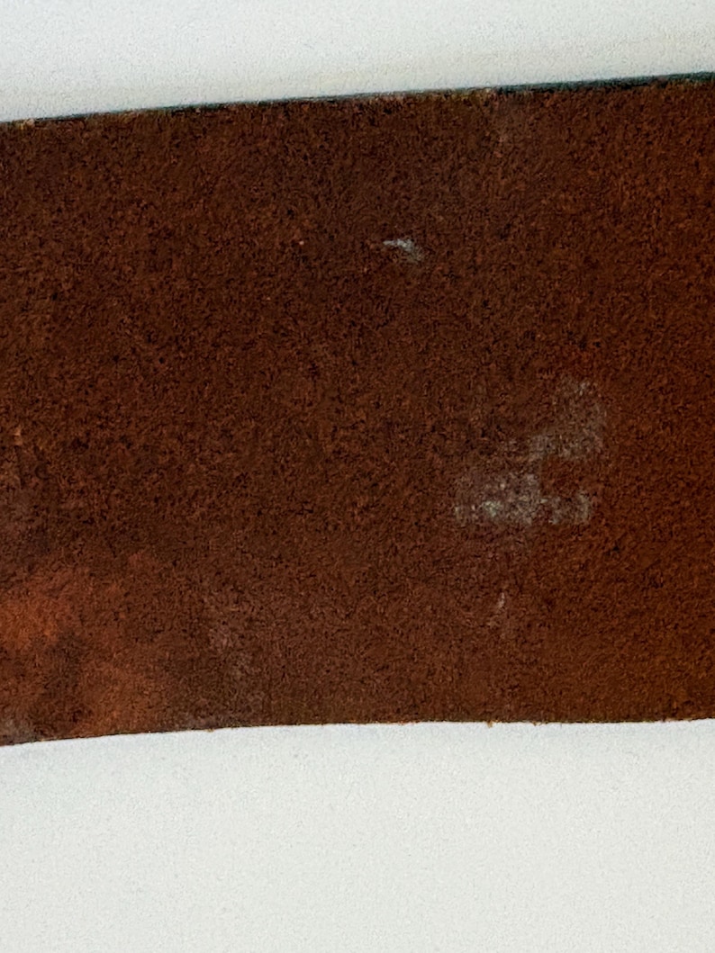 Puede incluir: Primer plano de un cintur&oacute;n de cuero marr&oacute;n. El cintur&oacute;n tiene una superficie texturizada con algunas manchas m&aacute;s claras. Los bordes del cintur&oacute;n son de color marr&oacute;n oscuro. El fondo es blanco.