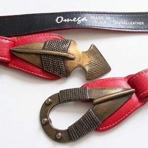 May include: A red leather belt with a decorative arrow-shaped buckle. The belt has a black leather strap with the text "Omega Made in U.S.A. 9444 Leather". The buckle is a bronze color.