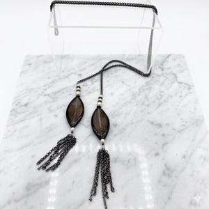 Puede incluir: Un collar plateado con dos colgantes en forma de hoja de color marrón oscuro. Cada colgante tiene una borla de cadena plateada y está adornado con pequeñas cuentas blancas. El collar se exhibe sobre una superficie de mármol.