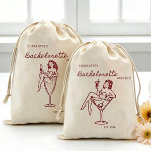 May include: Two off-white drawstring bags featuring a maroon graphic of a person sitting in a large martini glass. The text reads "CHARLOTTE'S Bachelorette" and "CHARLOTTE'S Bachelorette WEEKEND EST. 2026" in a maroon script and sans-serif font.