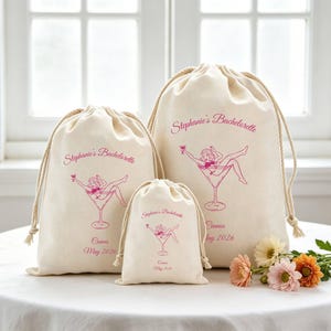 Puede incluir: Tres bolsas de algodón de color blanco roto con cordón de diferentes tamaños, cada una con un gráfico rosa de una persona en una copa de martini. El texto Stephanie's Bachelorette, Cannes y May 2026 aparece en cursiva en cada bolsa.