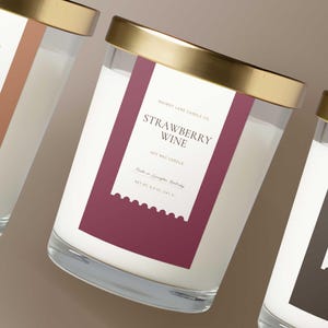 May include: Three glass candles with gold rims and labels. The labels read "Home & Hearth", "Strawberry Wine", and "Bonfire & Bourbon". Each candle is filled with white wax and has a label with the brand name and scent.