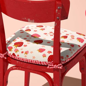 May include: A red wooden chair with a cushion featuring a strawberry pattern. The cushion has a white background with red strawberries and pink flowers. The chair has a distressed finish and a gingham border.