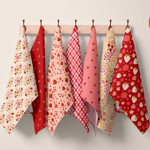 May include: A collection of six strawberry-themed kitchen towels hanging on a wooden rack. The towels feature various patterns, including strawberries, floral designs, and gingham checks, in shades of red, pink, and white. A circular sticker reads "Strawberry Delicious."