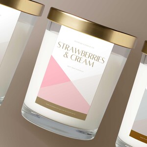 May include: Three glass jar candles with gold lids and labels. The labels feature geometric designs and text, including "Amber Noir," "Strawberries & Cream," and "Sand & Sea." The candles are filled with white wax.