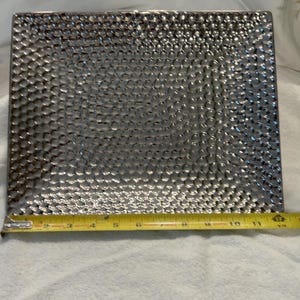 May include: A rectangular silver-toned metal tray with a hammered, dimpled texture across its entire surface. A yellow measuring tape is positioned along the bottom edge, indicating a width of approximately 12 inches for the tray.