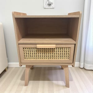 May include: A light brown wooden nightstand with a drawer featuring a woven rattan panel. The nightstand has a shelf above the drawer and is supported by four angled legs. The drawer has a light brown wooden handle.