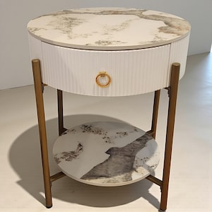 May include: A white, round side table with a marble-look top and lower shelf. The table has a single drawer with a gold ring pull. The legs are gold-colored and the drawer front has a vertical ribbed texture. The table is approximately 24 inches in diameter and 24 inches tall.
