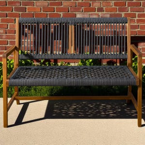 May include: A wooden outdoor bench with a woven seat and backrest. The seat and back are made of dark gray rope, and the frame is a light brown wood. The bench is set against a red brick wall.