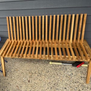 May include: A wooden bench with a slatted back and seat. The bench is made of light brown wood and has a simple, modern design. The back of the bench is angled, and the seat is flat. The bench is supported by four legs.