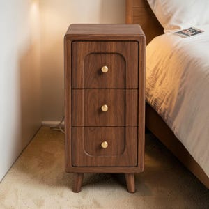 May include: A three-drawer wooden nightstand with a warm brown finish. Each drawer has a gold-colored knob. The nightstand has a rounded top and sits on four short legs. The nightstand is next to a bed with a white comforter.