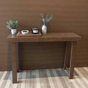 May include: A dark brown wooden console table with a rectangular top and four sturdy legs. The table is decorated with a small potted plant, a wooden tray with a bowl and candle, and a white vase with greenery.