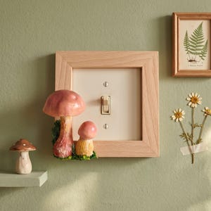 May include: A light wood light switch cover plate featuring two attached pink and yellow mushrooms with green moss accents. A separate small brown mushroom figurine sits on a shelf nearby, and a framed fern print hangs on the wall.