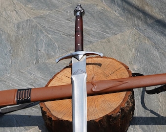May include: A silver sword with a brown leather scabbard. The sword is lying on a wooden log with the scabbard resting on top of it.