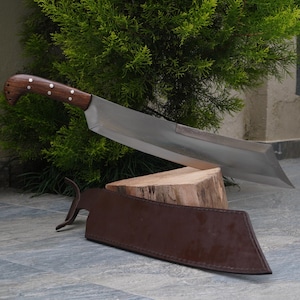 May include: A large, silver-colored knife with a wooden handle rests on a wooden block. The knife has a curved blade and a leather sheath.