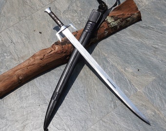 May include: A silver sword with a black leather scabbard and a wooden handle. The sword is lying on a wooden log with a leather strap attached to the scabbard.