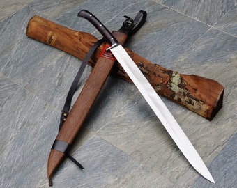 May include: A long, silver blade with a black handle is resting in a wooden scabbard with a leather strap. The scabbard is decorated with a red and white design.