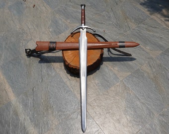 May include: A long, silver sword with a brown handle and crossguard, resting on a wooden log. A brown leather sheath with a black band and decorative accents lies beside it. The sword is displayed on a gray stone surface.