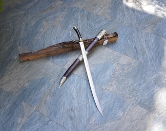 Pode incluir: Uma espada prateada com uma lâmina curva, punho preto e detalhes prateados ornamentados, repousando sobre um pedaço de madeira. A espada é acompanhada por uma bainha castanha escura com detalhes prateados. O fundo é uma superfície de pedra cinza texturizada.