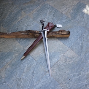 May include: A long, silver sword with a black handle and a brown leather sheath. The sword is resting on a piece of wood. The handle has a decorative design. The sheath is brown with a silver tip.