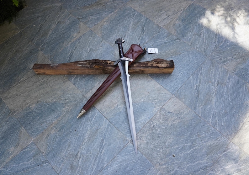 May include: A long, silver sword with a black handle and a brown leather sheath. The sword is resting on a piece of wood. The handle has a decorative design. The sheath is brown with a silver tip.